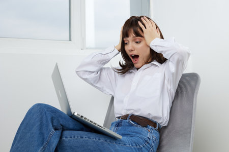A surprised young woman sits in a bright home office with a laptop on her lap, hands in her hair,...