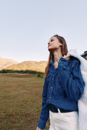 Woman outdoors in a grassy field near mountains, portrait wearing denim shirt and white jacket over shoulder, gazing at sky in a relaxed nature lifestyle scene.の写真素材