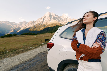 Woman with car beside mountains landscape for travel outdoors adventure roadtrip, standing confidently by a parked SUV on gravel road with scenic meadow and valley background at sunset.の写真素材