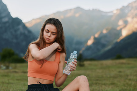 A young woman sits outdoors in a scenic meadow with mountains in the background, holding a water bottle. She wears an athletic top and looks relaxed, refreshed, and carefree.の写真素材