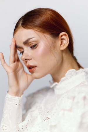 Young woman with red hair in a white blouse, expressing concern and contemplation against a soft, neutral background, capturing emotions of distress and reflectionの写真素材