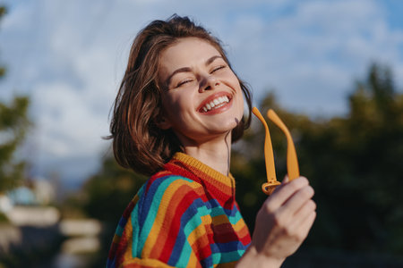 Woman smiling in a rainbow striped sweater holding yellow sunglasses outdoors, young adult female mid 20s portrait with short hair, joyful expression, casual outfit and natural sunlight lifestyleの写真素材