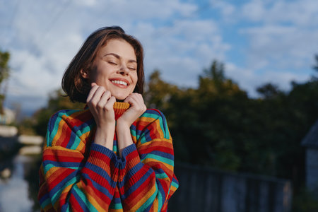 Young woman early 20s with short hair in a colorful striped sweater smiling with eyes closed, hugging collar outdoors by wooden railing in nature. Cozy joyful travel lifestyle.の写真素材