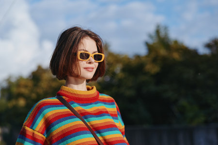 Woman in a colorful striped sweater and yellow sunglasses smiling outdoors, young adult with bob haircut wearing a casual outfit and crossbody bag, lifestyle travel portrait.の写真素材