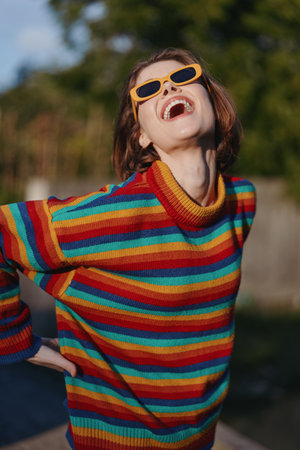 woman smiling striped sweater colorful sunglasses outdoors young adult joyful short hair casual outfit. Young woman in her 20s wearing a rainbow knit, leaning back and laughing in park sunlight forの写真素材