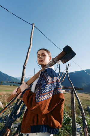 Woman with axe standing by wooden fence in mountains field, wearing patterned sweater on a rural farm. Portrait outdoors with rustic wood posts, clear blue sky and natural sunlight.の写真素材