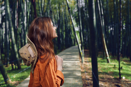 Woman in orange jacket hiking on a forest path through tall bamboo, portrait side view with hat on her back, enjoying nature walk and serene outdoor atmosphereの写真素材