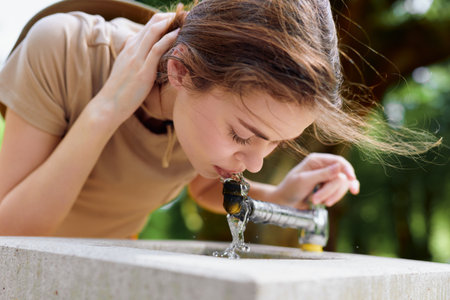 Woman drinking water from outdoor fountain, portrait closeup of young female bending to sip flowing tap water in park, casual summer scene with natural light and motionの写真素材