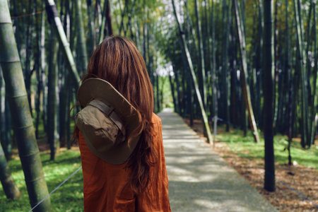 Woman with hat walking on a bamboo forest path, back to camera, nature scene with green trees and sunlight filtering along a peaceful trail for travel and relaxation.の写真素材