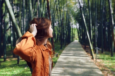 Woman on a path in a bamboo forest wearing an orange jacket and hat, walking and looking back along a sunlit trail, nature escape and peaceful outdoor portrait.の写真素材