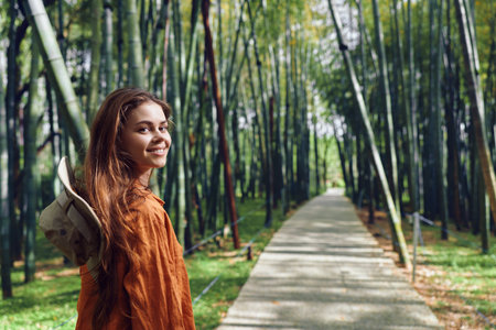 Woman walking on a paved path through bamboo forest, portrait of traveler with backpack and hat, smiling and looking back with a warm smile as sunlight lights the green outdoor sceneの写真素材