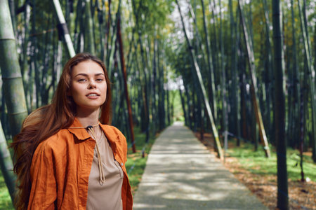 woman bamboo forest path portrait, traveler with backpack standing on paved walkway among tall bamboo stalks, wearing orange jacket in outdoor nature scene with calm, confident expressionの写真素材