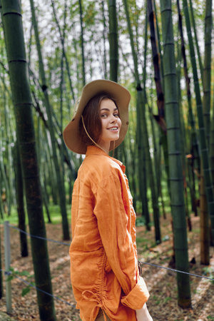 Woman in orange jacket and wide hat smiling in bamboo forest, portrait of a happy traveler surrounded by tall green bamboo and nature, casual outfit and relaxed outdoor walk.の写真素材