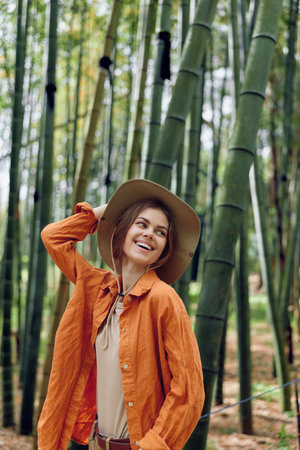 Woman smiling in hat among tall bamboo forest, nature portrait of a traveler wearing orange jacket and straw hat, candid outdoor moment showing eco tourism, adventure and joyful expression.の写真素材