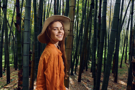 Woman in straw hat smiles in a bamboo forest portrait, wearing orange shirt during outdoor nature walk. Travel vibe, relaxed expression and green stalks creating a serene background.の写真素材