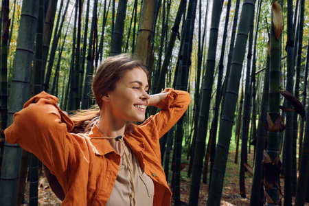 Woman in orange jacket smiling among tall bamboo forest, portrait of happy outdoor nature travel and relaxation enjoying sunlight and a peaceful green woodland scene.の写真素材