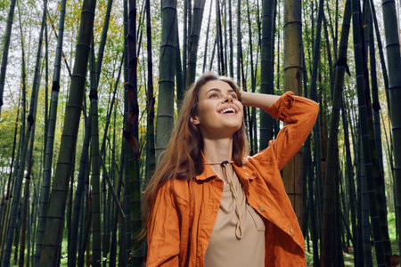 Woman in orange jacket stands in a bamboo forest smiling in a nature portrait, looking up with joy. Outdoors travel scene with relaxed lifestyle, sunlight and peaceful green woodland mood.の写真素材