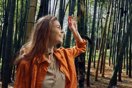 Woman in bamboo forest smiles and reaches up, portrait of a happy traveler in nature wearing an orange jacket, outdoor lifestyle and exploration with sunlight filtering through tall green stalks.の写真素材