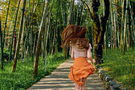 Woman running through a bamboo path in an orange skirt, back view capturing motion and flowing hair. Forest trail with sunlight and green nature evokes freedom and travel.の写真素材