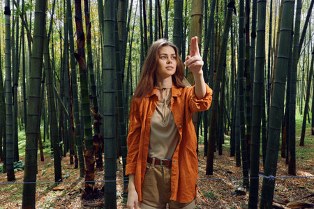 Woman bamboo forest pointing with raised hand in nature portrait, casual travel style among tall green trees. Young explorer standing in serene wood with thoughtful gaze.の写真素材