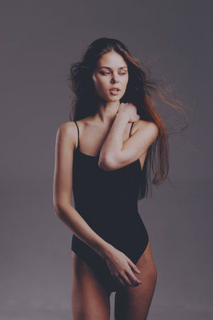 slender woman in black bodysuit posing with thoughtful expression and tousled hair on gray backgroundの写真素材