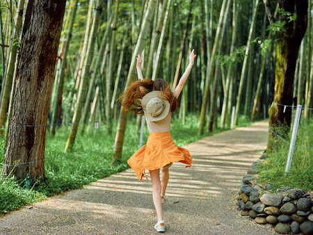 Woman in an orange dress and straw hat runs joyfully along a gravel path through a bamboo forest, back view in summer sunlight, carefree motion and outdoor nature escape.の写真素材