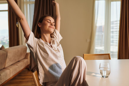 Woman in pajamas stretch at morning in a bright home, relaxed and smiling at a table with a glass of water, sunlight through the window in a cozy living room scene of calm wellbeing.の写真素材