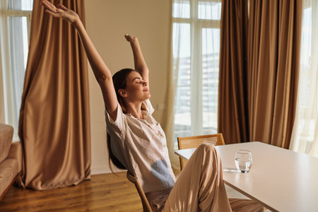 Woman relaxed stretching in morning at home, sitting on chair by table with glass of water, casual clothes and peaceful expression, natural light and cozy interiorの写真素材
