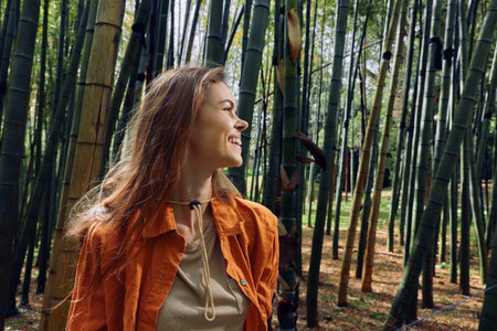 Woman portrait in bamboo forest smiling and looking sideways, nature outdoors with sunlight filtering through stalks, travel vibe, serene mood and casual jacket in green grove.の写真素材