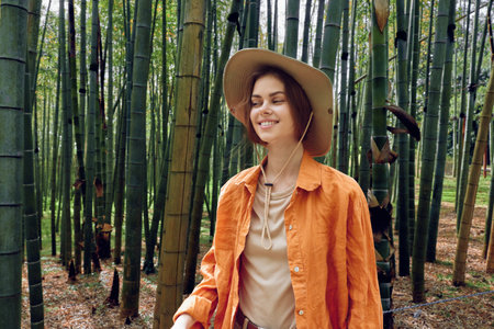 Woman walking in a bamboo forest wearing a hat and orange jacket, smile and portrait in nature. Outdoors travel and leisure scene among green trees conveying calm exploration.の写真素材
