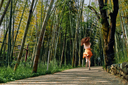 Woman running along a winding path through a dense bamboo forest, back view in a flowing orange dress. Motion blur, sunlight dappled trail, summer nature scene and carefree escape.の写真素材