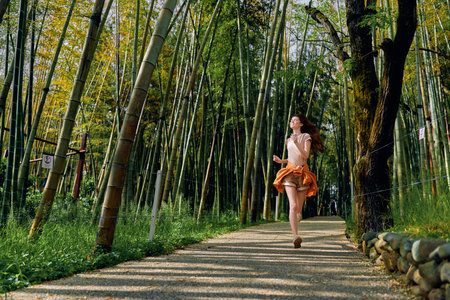 woman bamboo forest path running in a flowing dress, motion captured on a sunlit trail among tall green trees and canopy, outdoor nature scene with joyful movementの写真素材