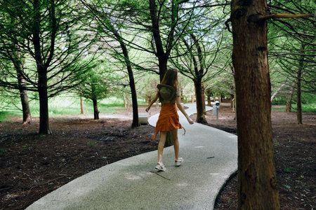 Woman walking path forest backpack dress trees trail nature outdoors summer sneakers solitude wander exploration with serene sunlight on curved pathway for relaxed outdoor journey.の写真素材