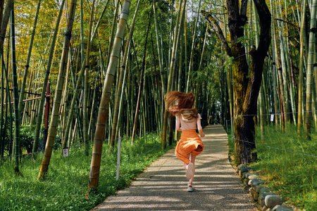 Girl running on a stone path through a dense bamboo forest wearing orange skirt, back view, motion and sunlight filtering through tall green stalks for outdoor nature walk.の写真素材