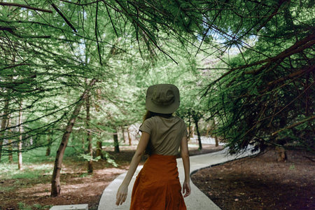 Woman with hat walking on paved path through green forest, trees and nature surrounding her. Back view of female in orange skirt exploring peaceful park trail.の写真素材