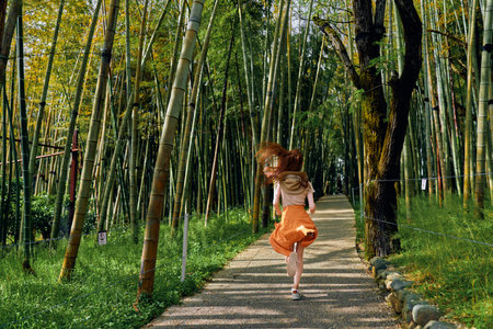 girl bamboo path running in a green forest, back view of a barefoot child in orange skirt on a sunlit trail, motion and freedom in nature during summer outdoors exploration.の写真素材