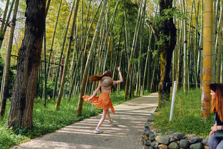 Woman in a hat and orange dress walking and twirl on a gravel path through a bamboo forest in summer nature, joyful movement and carefree exploration under sunlight and leafy trees.の写真素材