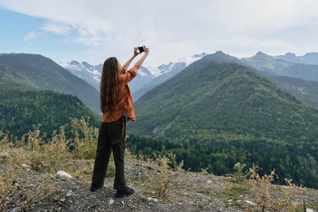 Woman stands on a rocky overlook taking a photograph of the vast mountain landscape with forested valleys and distant peaks under a bright blue skyの写真素材