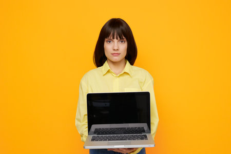 Young woman stands upfront with a laptop extended toward the camera against a bright orange background, presenting a clean, modern tech inspired portrait.の写真素材