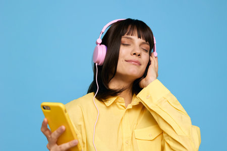 A person wearing pink headphones and a bright yellow shirt listens to music on a smartphone, smiling softly against a solid blue studio backdrop.の写真素材