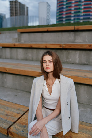 Young businesswoman in white suit sitting on urban wooden steps with thoughtful expression, modern cityscape backgroundの写真素材