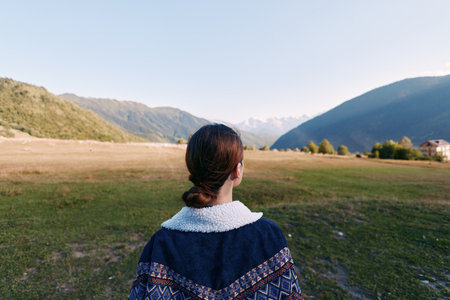 Woman back overlooking mountain landscape and meadow in a valley, outdoors travel scene with calm nature, rural field and distant hills for peaceful exploration and adventure.の写真素材