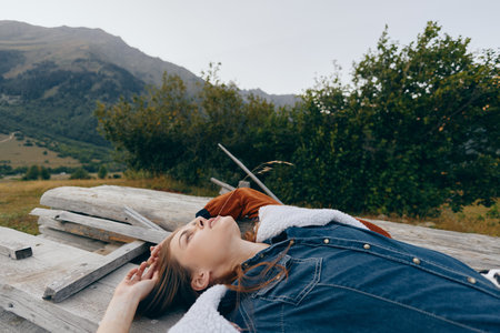 Woman lying on wooden bench in nature with mountains background, relaxed portrait in denim jacket outdoors, cozy layers and peaceful relaxation during countryside escape.の写真素材