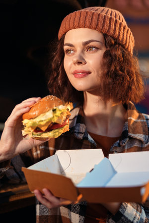 Woman holding burger and takeout box, smiling in beanie and plaid coat with streetfood vibe, authenticity and candid lifestyle captured in golden hour glow, emotional storytelling.の写真素材
