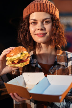 Woman holding burger and takeaway box, smile and beanie with plaid shirt in evening sunlight and golden hour glow. Authenticity, candid lifestyle and emotional storytelling evoke mindful living.の写真素材