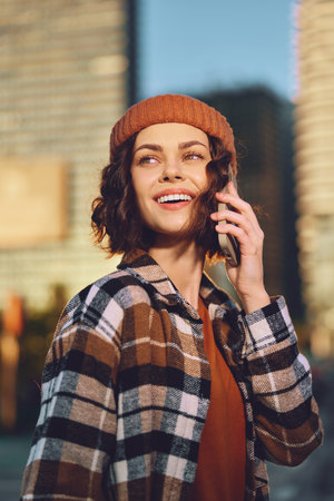 Woman on phone smiles in city portrait wearing beanie and plaid jacket, outdoor sunlight casting golden hour glow. Authenticity and candid lifestyle captured with mindful living vibe.の写真素材