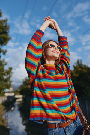 Young woman in a colorful striped sweater and yellow sunglasses smiling and stretching arms outdoors under blue sky, casual outfit, joyful lifestyle and urban nature backdrop.の写真素材