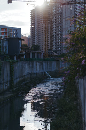 Urban canal with concrete canal and highrise buildings framed by a construction crane at sunset reflection, water stream from a drainage pipe and riverside vegetation in moody twilight.の写真素材