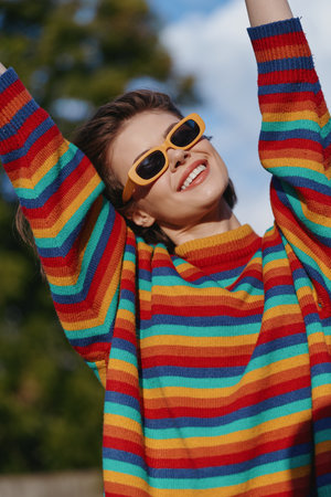 woman striped sweater sunglasses smiling young adult outdoors colorful sweater casual outfit; joyful young woman with yellow shades raising arms, sunlit lifestyle portrait for fashion and travel.の写真素材