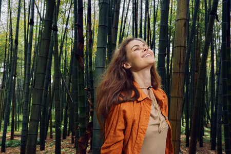 Woman in a bamboo forest smiles while looking up, enjoying nature outdoors with sunlight filtering through tall green stalks; portrait of happy relaxed adult in orange jacket.の写真素材